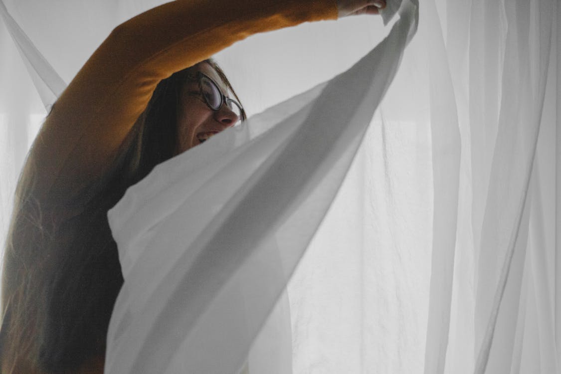 Cheerful young lady having fun in bedroom while hiding behind curtains