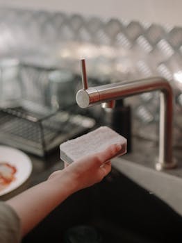 A hand holding a sponge by a kitchen sink, ready for cleaning.