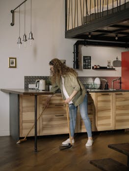 Young woman cleaning a modern loft kitchen area with a broom, demonstrating home maintenance.