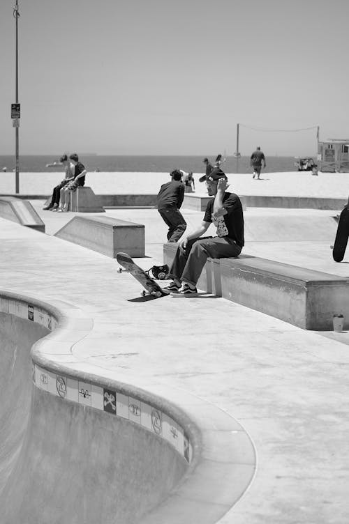 Side view of black and white anonymous male skateboarder resting on bench during outdoor training in skate park on sunny day