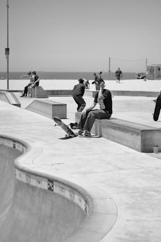 Side view of black and white anonymous male skateboarder resting on bench during outdoor training in skate park on sunny day