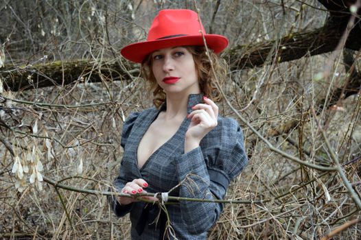 Calm young female with ginger hair in elegant dress and red hat standing in forest amidst leafless branches of fallen tree and looking away