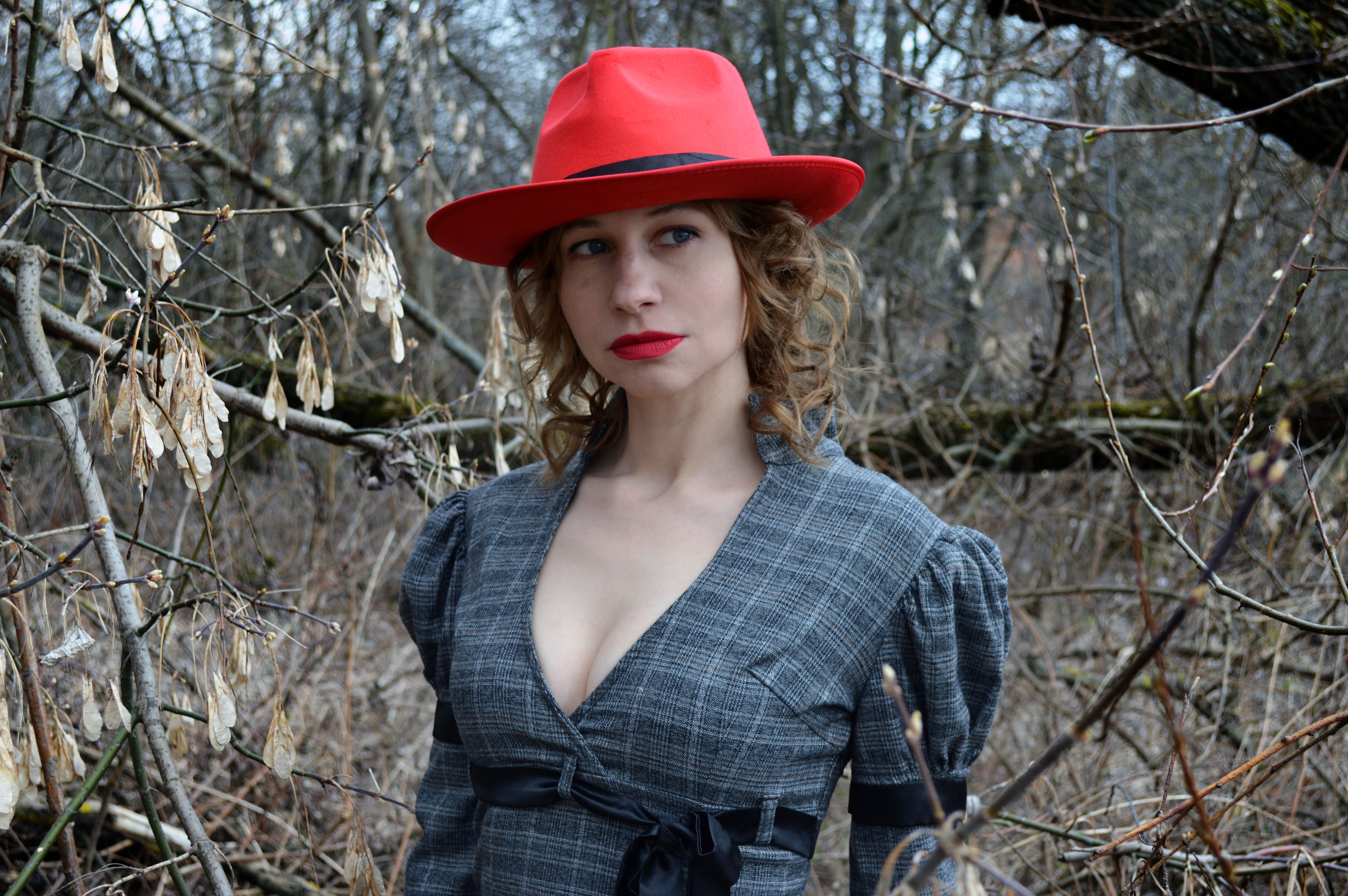 Young female with red lips and hat in checkered dress with decollete relaxing in park near leafless trees and looking away