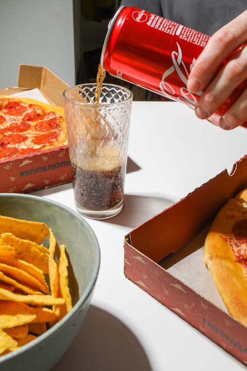 Person Pouring Coca-Cola On Glass Beside Snacks