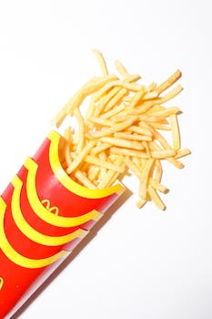 Golden crispy French fries spilling from a distinct red container on a white backdrop.