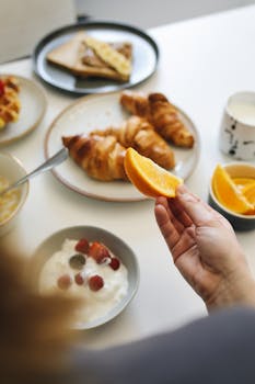 Hand holding an orange slice over a breakfast spread with croissants and yogurt.