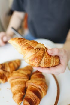 Close-up of a fresh croissant being held, with more pastries in the background, highlighting texture and freshness.