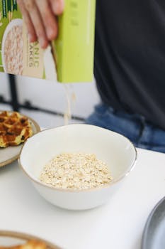 Person pouring organic cereal flakes into a breakfast bowl with waffles nearby.