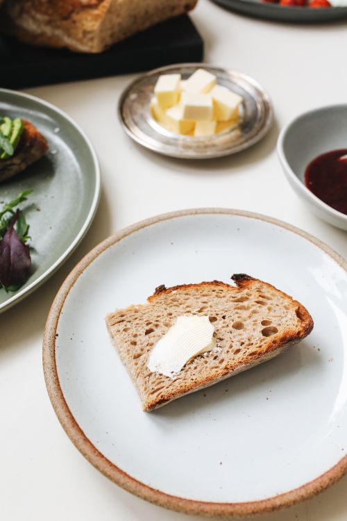 Free Slice of bread with butter on ceramic plate, surrounded by gourmet breakfast items. Stock Photo