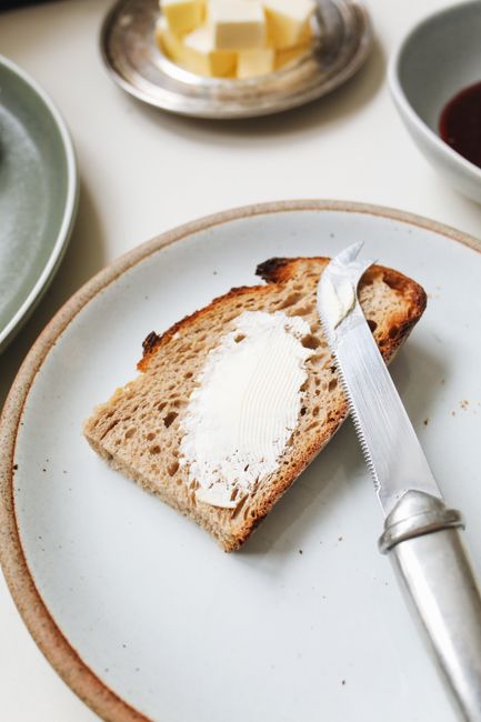 A block of pale yellow homemade dairy-free butter on a small dish, partially spread on a slice of toast with a butter knife resting beside it.