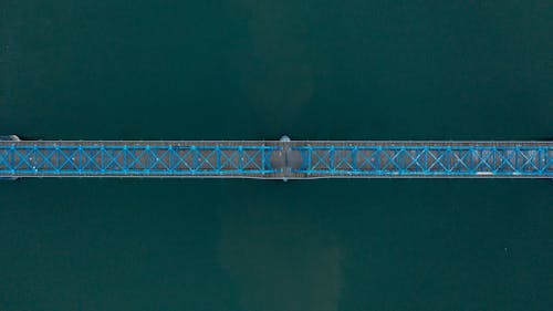 Drone view of distant modern boat floating under suspension bridge over calm river during daytime