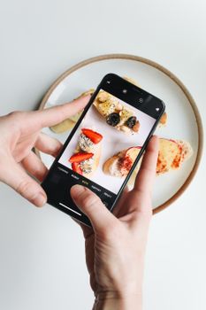 Close-up of hands photographing a plate of gourmet cheese topped snacks with a smartphone.