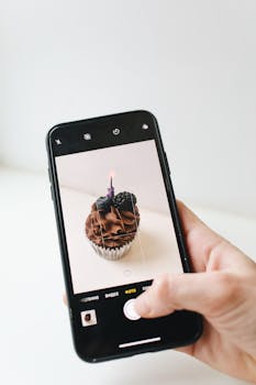 Hand holding smartphone capturing a chocolate cupcake with a lit candle.