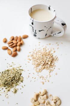 Overhead view of almonds, oats, hemp seeds, cashews, and milk in a modern mug on white surface.