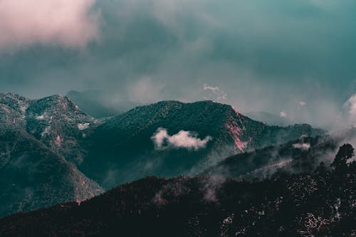 Majestic landscape of mountains covered with trees with snowy peaks under floating clouds on foggy day