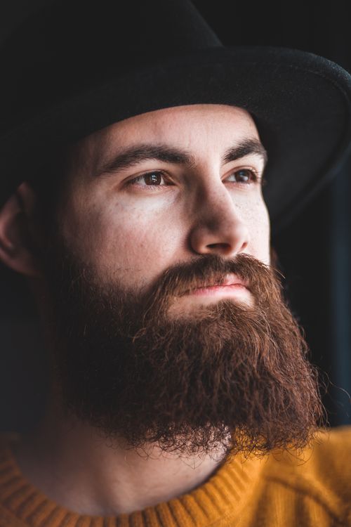 Free Serious young bearded male in warm sweater and trendy hat standing near window during daytime and looking away thoughtfully Stock Photo