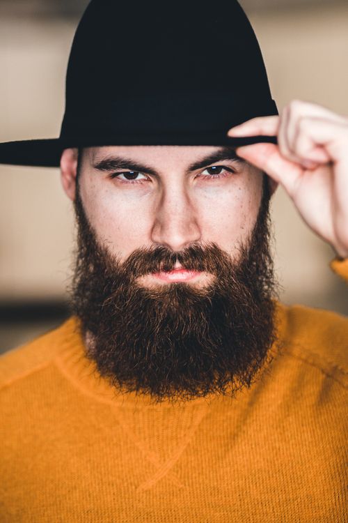 Free Portrait of a bearded man in a fedora and sweater with an intense gaze, embodying modern fashion. Stock Photo
