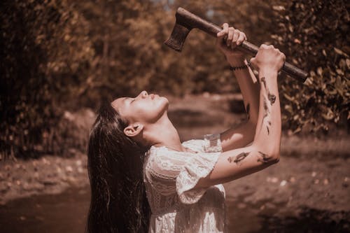 Side view of young melancholic ethnic female with long dark hair and closed eyes standing in autumn forest and holding axe over face