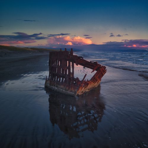 Free Rusty shipwreck remains at sunset on a serene Oregon beach, reflecting dramatic cloudscape. Stock Photo