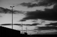 Low angle of black and white distant car with headlights riding on dark rural road near fences and streetlights against cloudy sky in evening