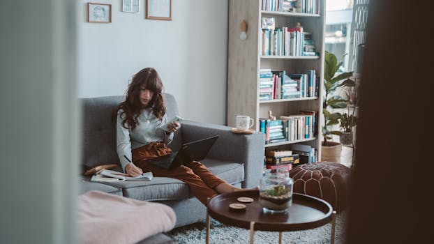 Woman seated on a sofa working remotely with a laptop and phone in a cozy home setting.