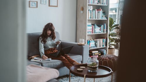 Free Woman seated on a sofa working remotely with a laptop and phone in a cozy home setting. Stock Photo