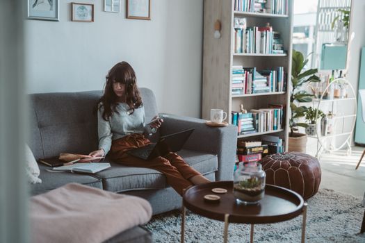 A woman working remotely at home on her laptop, seated on a comfortable sofa surrounded by bookshelves.