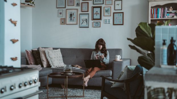 A woman multitasks on a sofa using a laptop and smartphone in a cozy home interior.