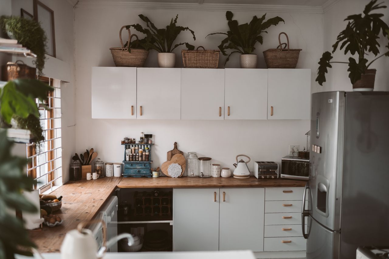 Cozy L-shaped kitchen with flat-front white cabinets, wood counters, and plants on top of the cabinets