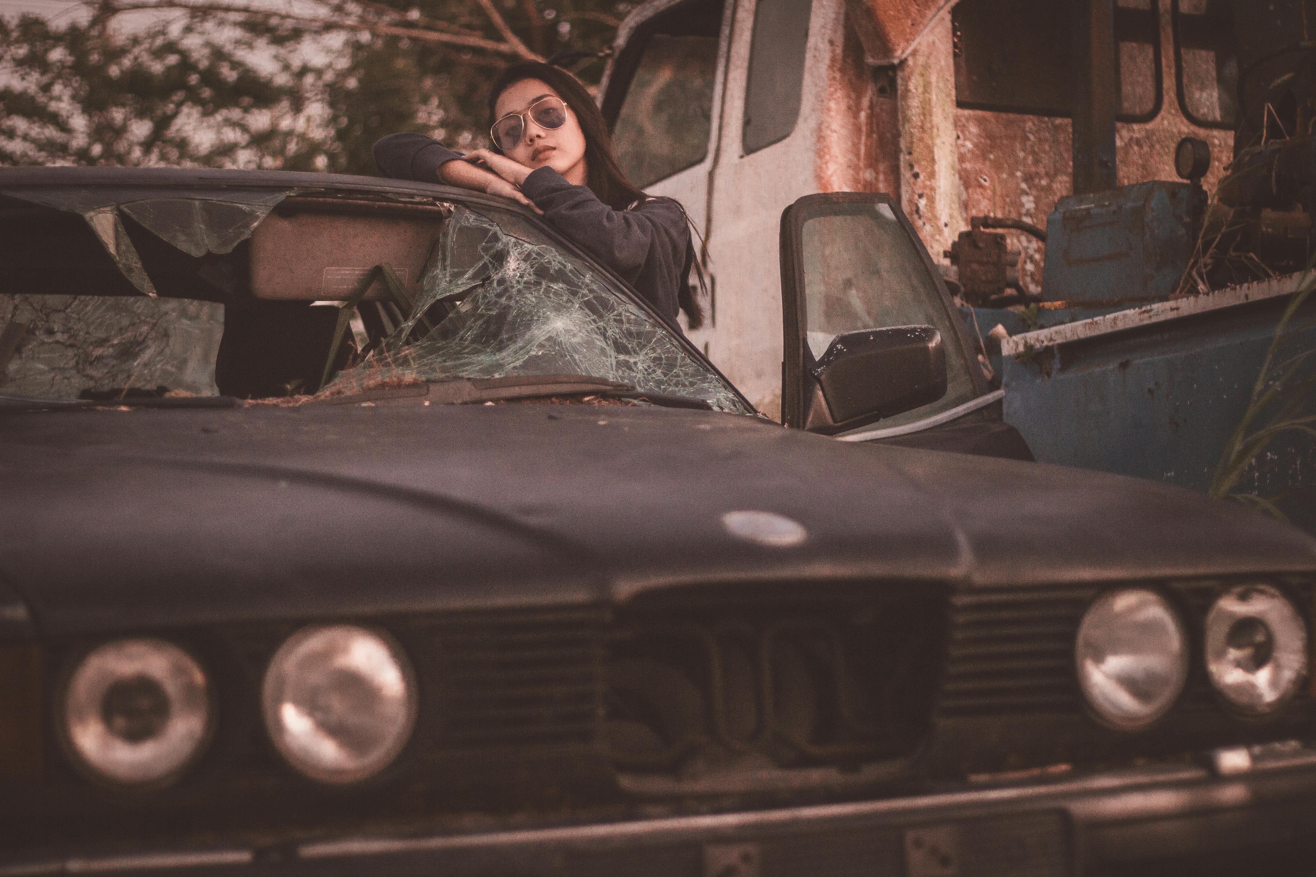 A woman in sunglasses leaning on a broken car in a rustic setting.