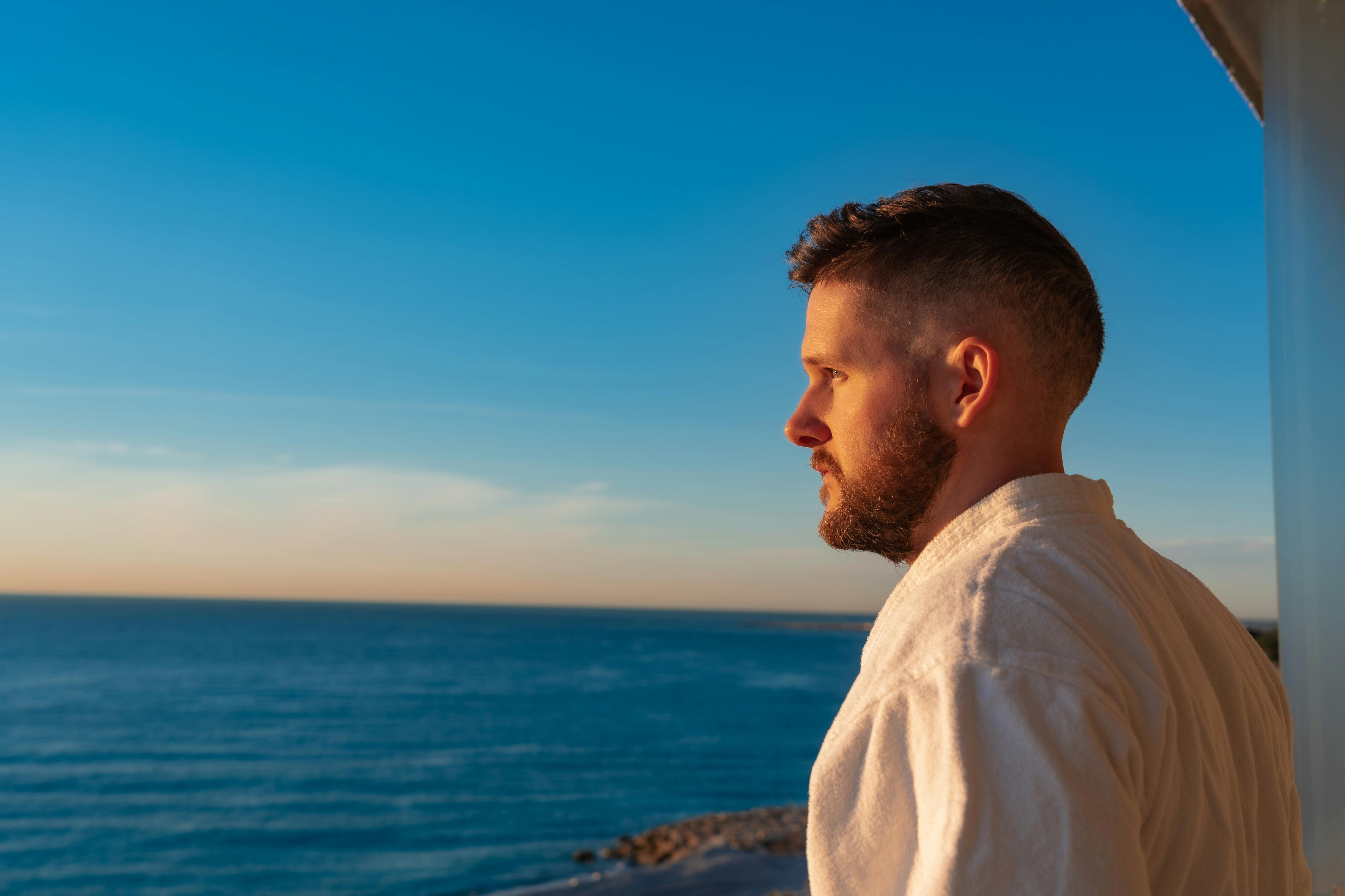 A man with a beard gazes at the ocean during sunset, wearing a white robe, reflecting tranquility.