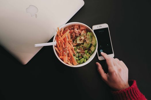 A bowl of fresh salad with avocado, carrots, edamame, and quinoa on a desk beside a smartphone and laptop.