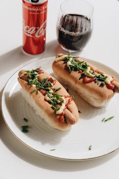 Two hot dogs with toppings next to a soda can and glass on a white table.