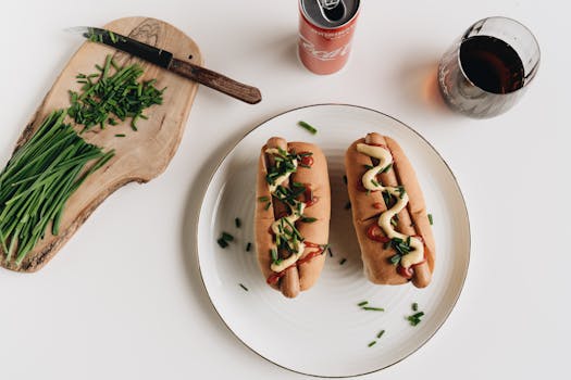 Top-down view of gourmet hot dogs with chives, served with soda on a wooden board.
