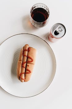 Delicious hot dog with ketchup and a refreshing soda served on a white plate.