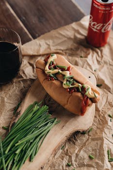 Rustic hot dog topped with chives, ketchup, and mustard, served on wooden board with a drink.