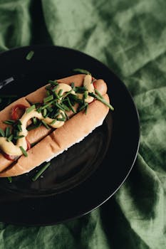 Artfully arranged hot dog with mustard and chives on a black plate, perfect for gourmet street food lovers.