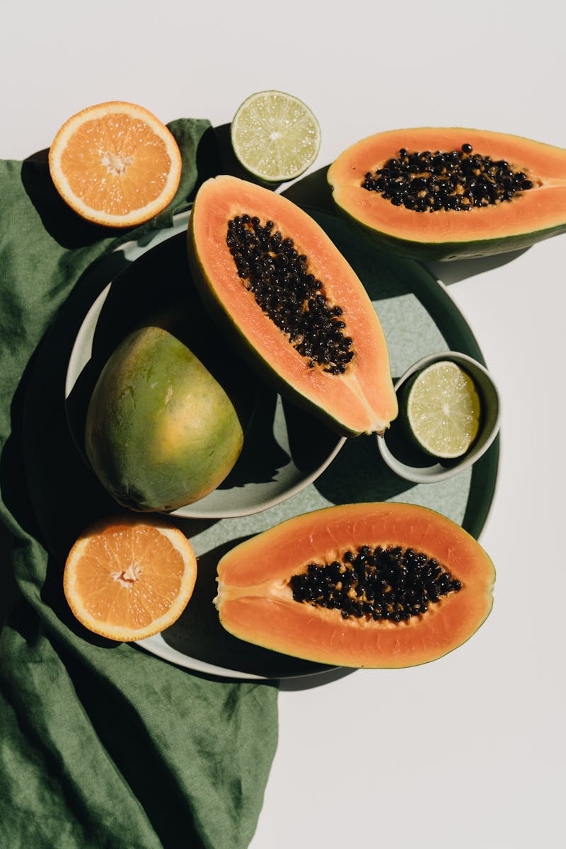 Top view of halves of ripe papaya together with oranges and limes placed on green round dishes and green fabric on white background