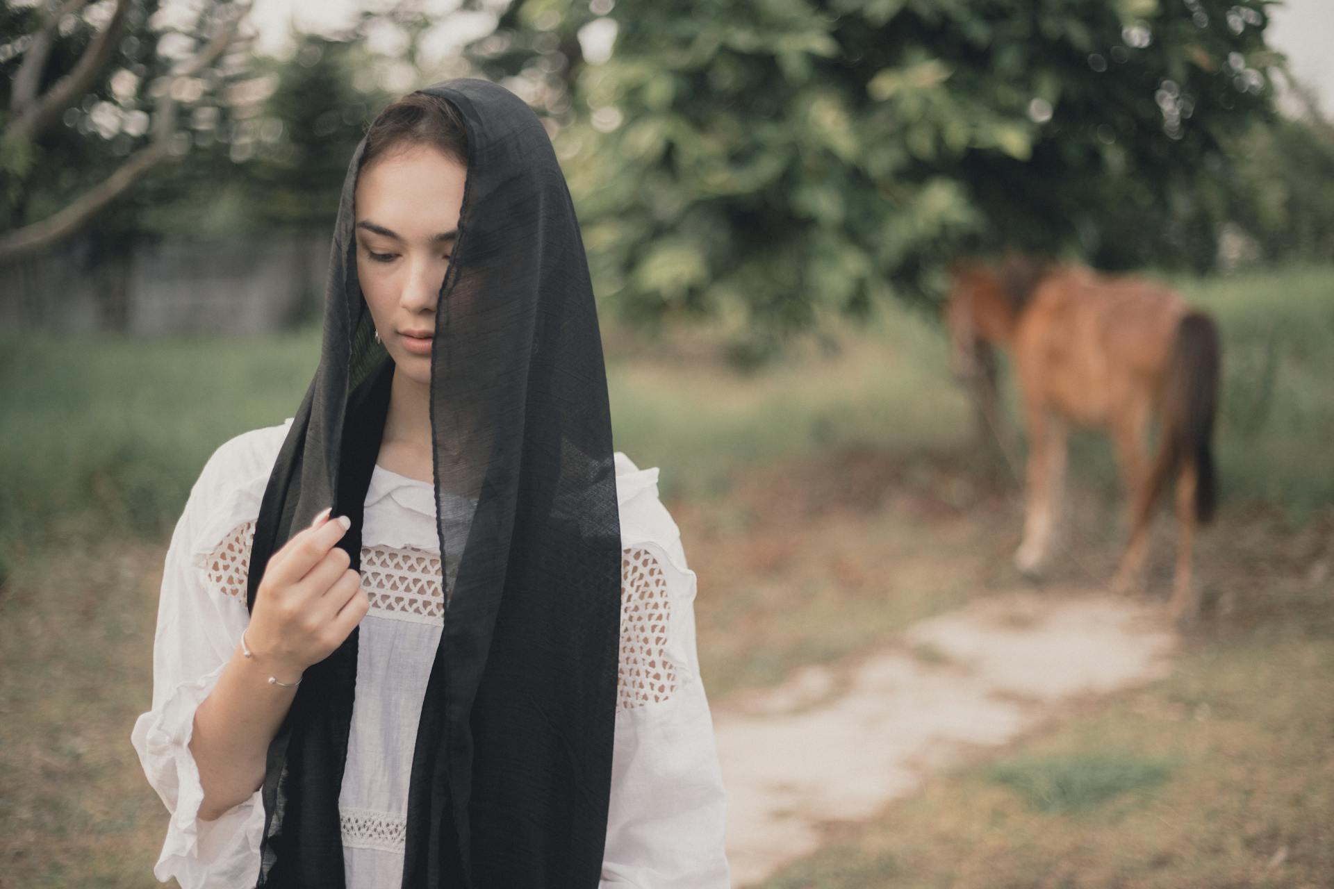 Woman in black headscarf and white dress in a calm rural setting with a horse in Cavite City, Philippines.