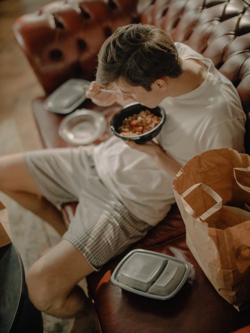 A relaxed young man eating takeout food on a leather couch in a cozy home setting. A relaxed young man eating takeout food on a leather couch in a cozy home setting.