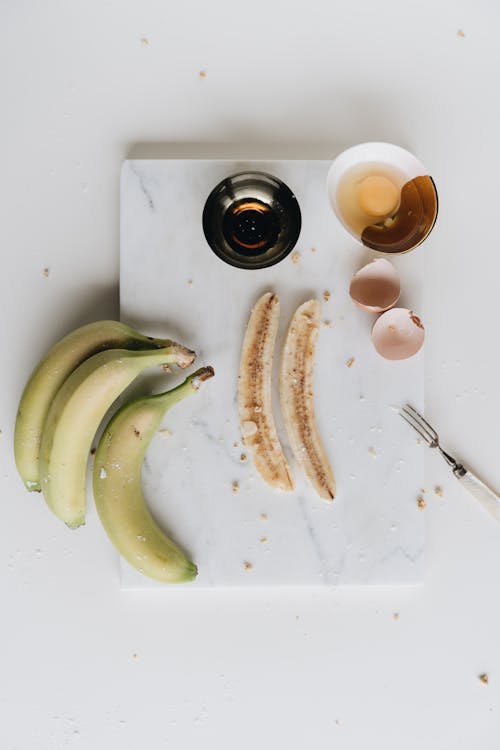 Free Top view composition of cut ripe bananas and bowl of raw egg on white marble board decorated with broken eggshell and fork Stock Photo