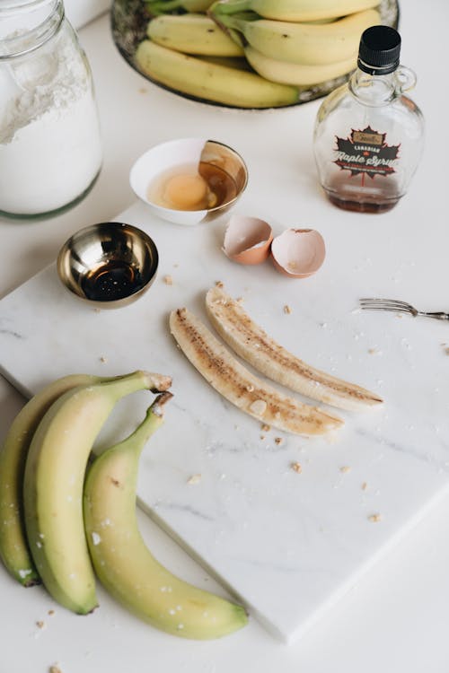 From above composition of healthy organic cut banana under syrup on white marble board with bowl of raw egg decorated with eggshell