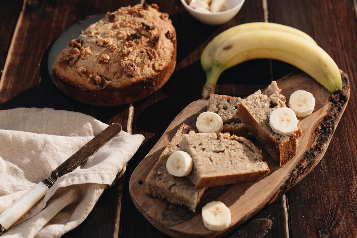 Sliced banana bread on a wooden board with banana rounds, a walnut-topped loaf and bananas