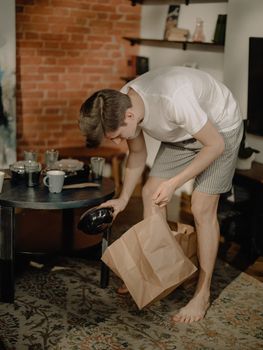 Young man cleaning up his living room with a paper bag, in a cozy home setting.