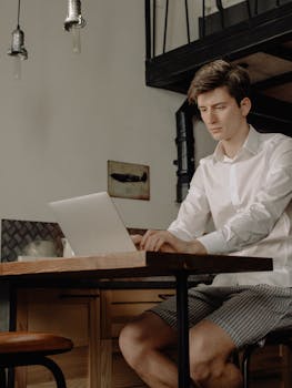 Young man in casual attire working on a laptop at home with a modern interior.