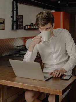 Man in a kitchen working on a laptop while sipping coffee, embodying the home office lifestyle.