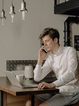 Young man multitasking with laptop and phone in modern home office.