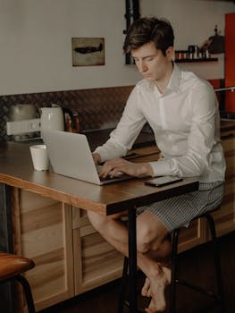 A man works on his laptop at home in a cozy kitchen setting, wearing casual attire.