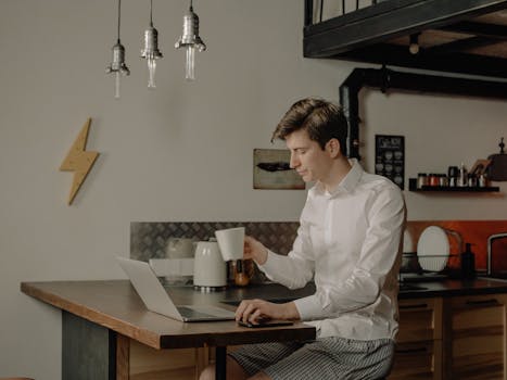 A young man in a modern kitchen using a laptop for remote work, enjoying a coffee break.