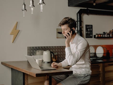Young man multitasking at home office with laptop and phone, embodying work-from-home lifestyle.